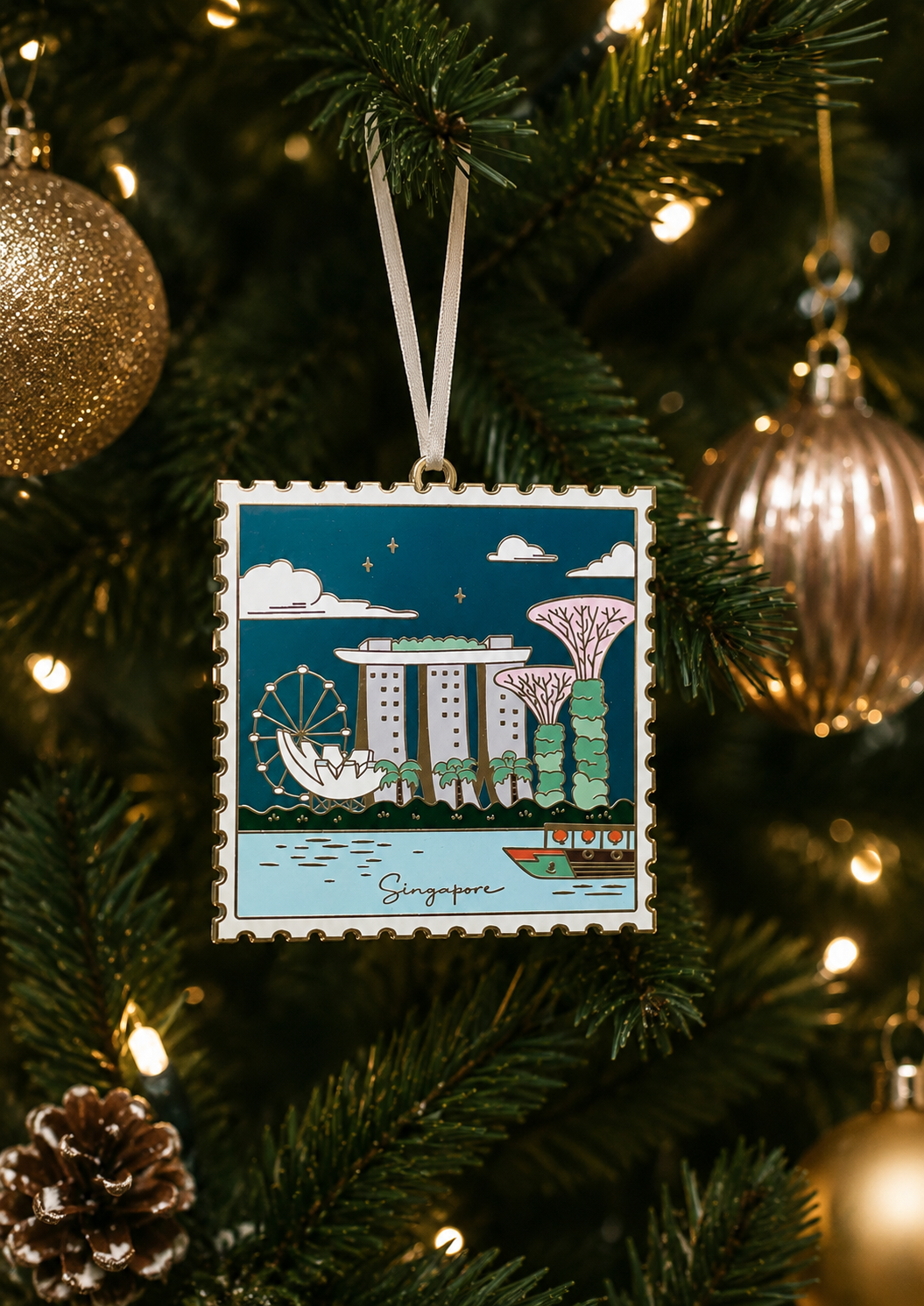 A square Christmas ornament featuring an illustrated view of Singapore, hanging from a green Christmas tree with warm lights and gold ornaments in the background.