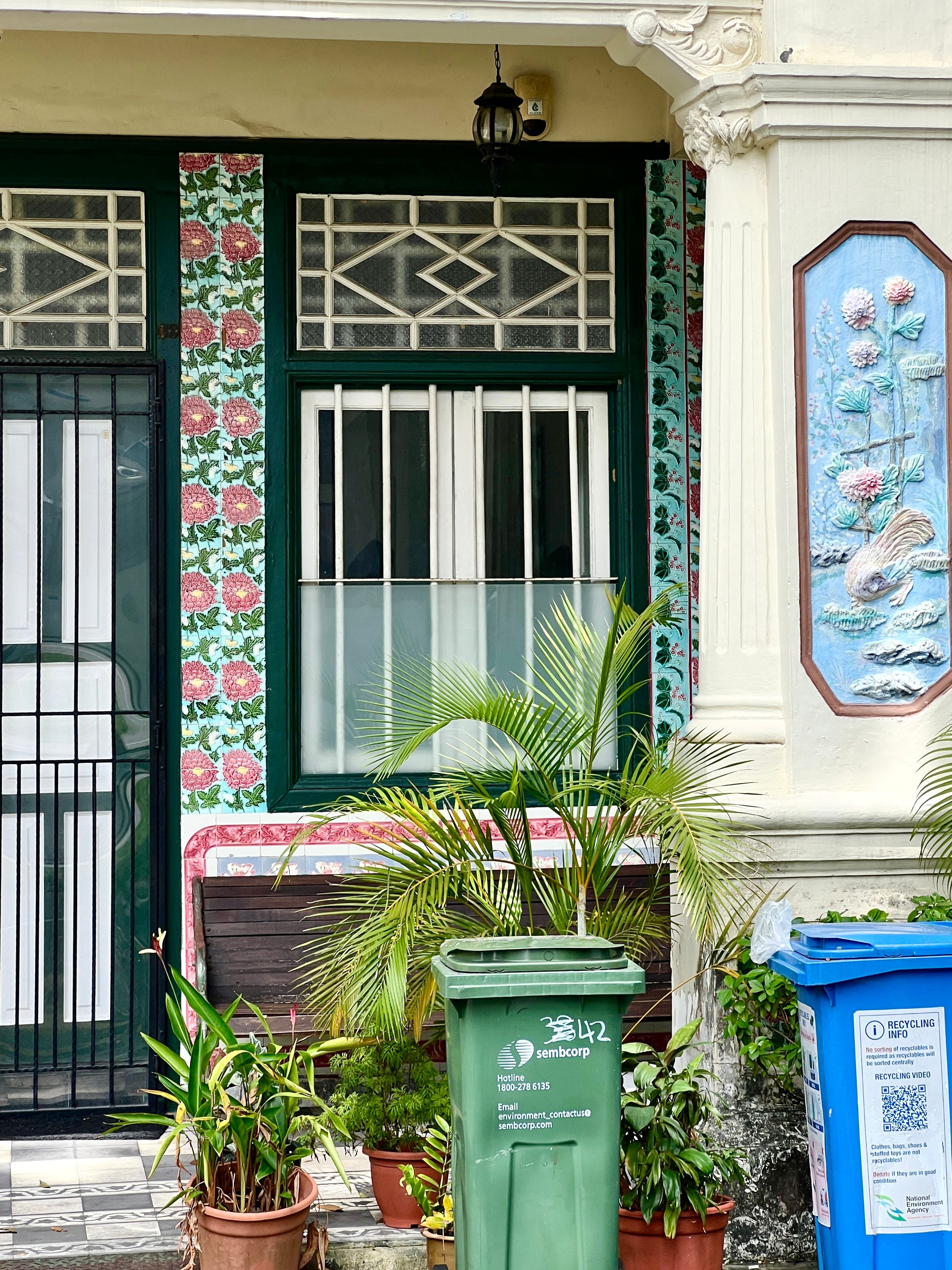 Decorative entrance with floral patterns, green and blue trash bins, and potted plants.